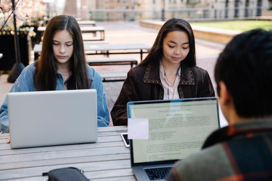 Two students focus on studying with laptops at an outdoor university setting, promoting technology and education.